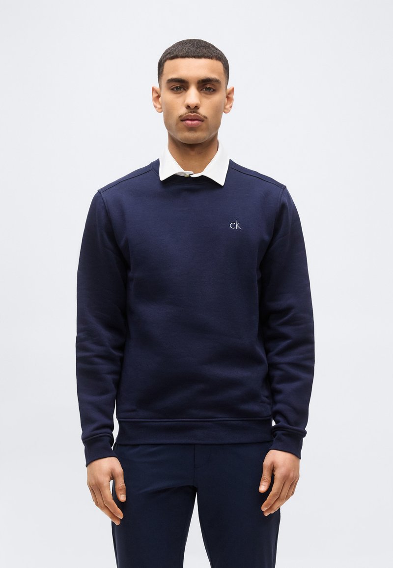 Άνδρας που φοράει μπλε σκούρο φούτερ με λευκό πουκάμισο από μέσα, στέκεται μπροστά σε απλό ανοιχτό φόντο, κοιτάζοντας μπροστά.