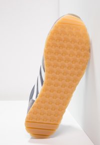 Sole of gray Adidas sneaker with white stripes, honeycomb-patterned light brown rubber outsole, angled against a white corner wall.