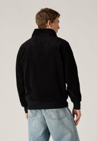 Μαύρο φλις πουλόβερ με ψηλό γιακά, μαλακή υφή και ριμπ μανσέτες. Φορεμένο με ανοιχτό μπλε τζιν σε άνετη γραμμή. Απλός σχεδιασμός, χωρίς ορατά λογότυπα.