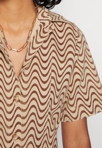 Camisa de botão feita de tecido bege com um padrão ondulado castanho. Apresenta colarinho, mangas curtas e botões visíveis à frente.