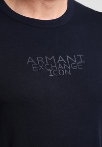 Černé tričko s kulatým výstřihem s nápisem "ARMANI EXCHANGE ICON" vytištěným světle šedými písmeny na hrudi.