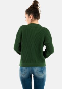 Pull vert en tricot avec une finition texturée, col montant et poignets côtelés, associé à un jean bleu ajusté avec un design unique de poche arrière.
