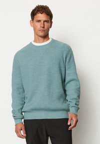 Hellblauer Pullover mit gerippter Textur, rundem Ausschnitt und langen Ärmeln. Über einem weißen Hemd getragen, kombiniert mit schwarzen Hosen.