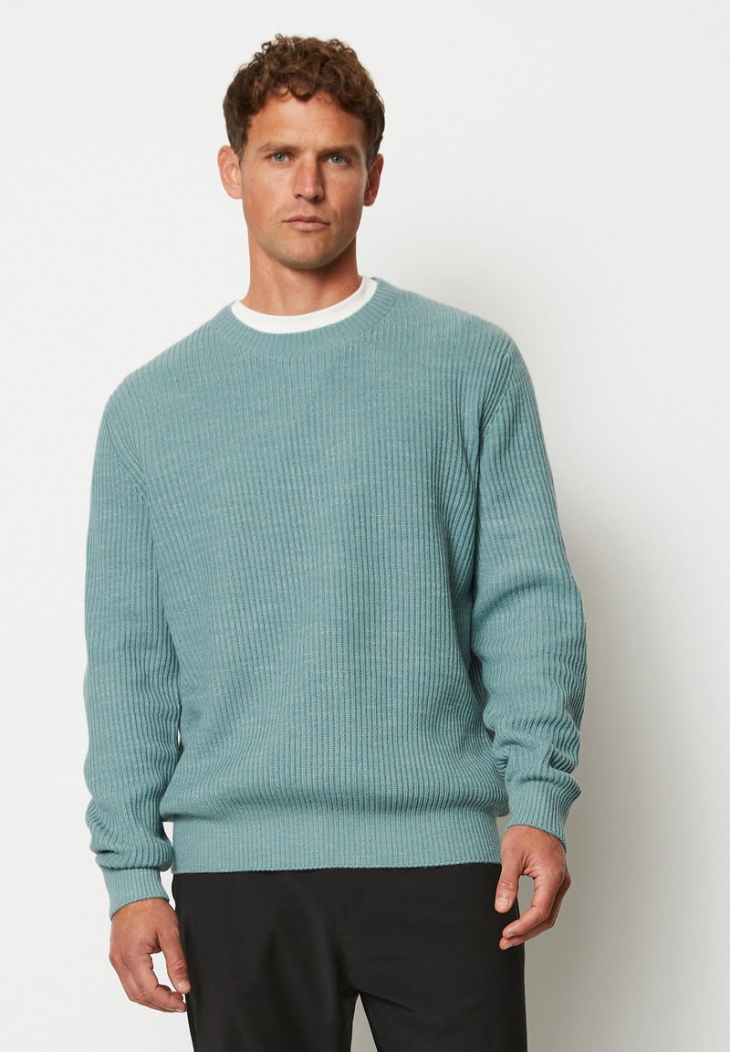 Hellblauer Pullover mit gerippter Textur, rundem Ausschnitt und langen Ärmeln. Über einem weißen Hemd getragen, kombiniert mit schwarzen Hosen.