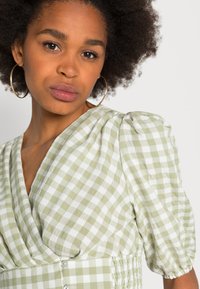 Vestido em padrão de xadrez verde e branco com mangas bufantes, decote cruzado e detalhes em botão na cintura. Tecido de algodão com uma textura suave.