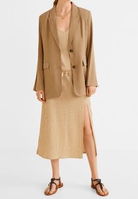 Kvinde iført en tan blazer over en beige og hvid vertikalt stribet midikjole med en sideslids og brune lædersandaler.