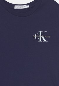 Tummansininen puuvillapaita, jossa on pyöreä pääntie ja lyhyet hihat. Paidassa on valkoinen "CK"-logo ja "CALVIN KLEIN JEANS" -teksti kullassa.