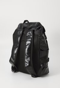 Zaino nero realizzato con materiali opachi e lucidi, caratterizzato da pannelli laterali in rete, spallacci regolabili e tasche con zip sulla parte anteriore.
