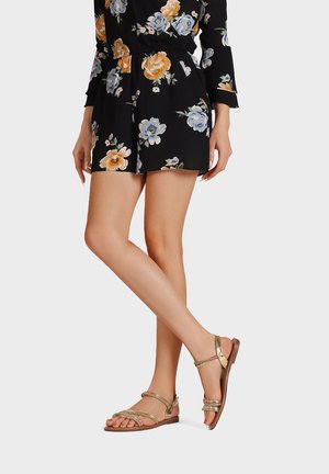 Donna che indossa pantaloncini neri floreali e sandali piatti dorati con cinturini, in piedi con le gambe incrociate e mani rilassate ai lati.