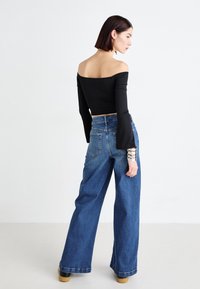 Black off-the-shoulder top, long sleeves, paired with high-waisted blue wide-leg jeans. Model wears silver cuff bracelet and brown platform shoes.