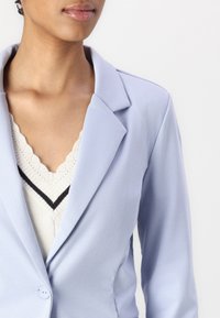 Ljusblå blazer med spetsig krage, strukturerade axlar och en slät textur, lagd över en krämfärgad stickad topp med scallopkant.