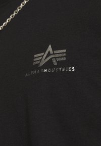 Camiseta negra de tejido suave con un logo metálico plateado que incluye el texto "ALPHA INDUSTRIES" y un diseño geométrico.
