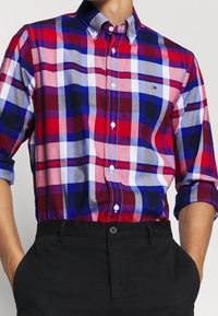 Camisa abotonada a cuadros en rojo, azul y gris, con patrones de cuadros contrastantes y un pequeño logo en el pecho. Tela de algodón, manga larga.