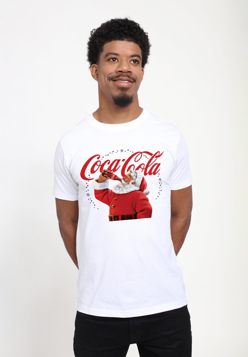Bílý bavlněný tričko s grafikou Santa Clause v červeném obleku, jak pije z láhve Coca-Cola, s červeným písmem a hvězdami.