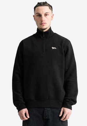 Mann mit geflochtenem Haar, der einen schwarzen Viertel-Reißverschluss-Sweatshirt mit kleinem weißem Tigerlogo auf der Brust trägt, steht vor einem schlichten hellen Hintergrund.