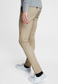 Chinos beige con un corte ajustado y pliegues sutiles, combinados con zapatillas blancas. La tela es suave con un ligero brillo.
