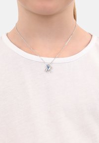 Collana d'argento con una figura montata su un cavallo, con un accento blu. La catena ha un design delicato, indossata sopra una camicia bianca.