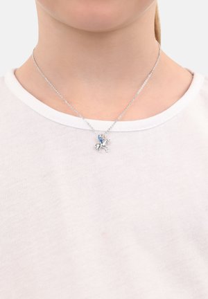 Silberne Halskette mit einer auf einem Pferd montierten Figur, die einen blauen Akzent aufweist. Die Kette hat ein zartes Design und wird über einem weißen Hemd getragen.