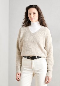 Beige strikket genser med V-hals og løs passform, med et teksturert mønster, kombinert med hvite høytlivsbukser og et svart belte.