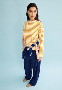 Camicia a maniche lunghe a righe arancione e bianca; abbinata a pantaloni a gamba larga blu navy. Un wrap a strisce marrone chiaro e blu attorno alla vita.