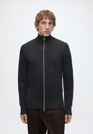 Jeune homme aux cheveux bouclés portant un pull zippé sombre et un pantalon foncé, debout devant un fond clair uni.