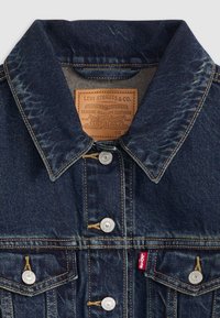 Casaco de ganga azul escuro com botões prateados, gola clássica e detalhes costurados. Possui uma etiqueta de couro castanho com informações da marca.