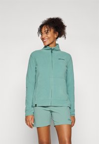 Veste en polaire vert clair avec fermeture éclair sur le devant, col montant et grandes poches latérales. Associée à un short assorti avec un logo.