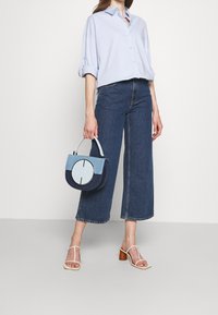 Woman wearing light blue button-up shirt and wide-leg cropped jeans, holding a blue and denim circular handbag, wearing white heeled sandals.