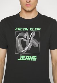 Camiseta negra de algodón con un gráfico en blanco y verde neón que dice "CK" y el texto "CALVIN KLEIN JEANS" debajo, de mangas cortas y cuello redondo.