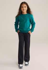 Felpa turchese con un design a forma di V decorato da borchie, abbinata a pantaloni neri a gamba larga. L'outfit è completato da sneakers bianche e nere.