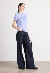 Violetinio medvilninio marškinėlių su užrašu "TOMMY JEANS" tamsiu spaudiniu, derinamas su plačiomis tamsiomis denimo kelnėmis ir juodomis plokščiomis bateliais. Aksesuarai: mažas juodas krepšys.