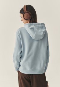 Világoskék kapucnis pulóver húzózsinórral, bordázott mandzsettával és szegéllyel. Barna, szőrös fülvédő medve mintával a fülvédőn.