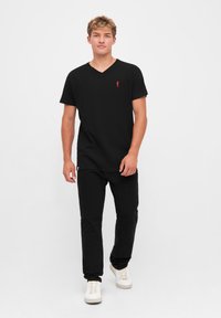 Jeune homme aux cheveux bouclés portant un T-shirt noir à col en V, un pantalon noir et des baskets blanches, se tenant contre un fond blanc.