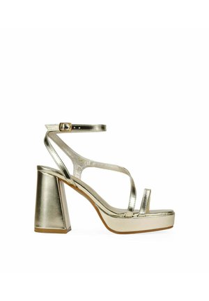 Sandalia metálica dorada de tacón alto con tacón grueso, correa en el tobillo y punta abierta sobre fondo blanco.
