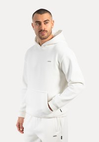 Bílá mikina s hladkým materiálem, přední kapsou, žebrovanými manžetami a malou nášivkou na hrudi. Nastavitelná šňůrka na kapuci.