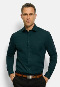 Donkergroen shirt met lange mouwen, knoopsluiting aan de voorkant, enkel borstzakje en een gladde textuur, gecombineerd met een bruine riem en donkere broek.