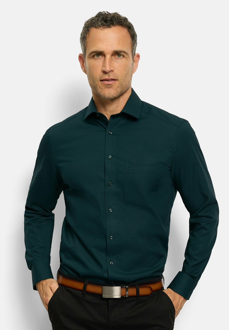 Donkergroen shirt met lange mouwen, knoopsluiting aan de voorkant, enkel borstzakje en een gladde textuur, gecombineerd met een bruine riem en donkere broek.