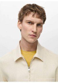 Beige zip-up skjorta med krage, med en silvrig dragkedja och en ficka, bärs över en gul t-shirt med rund hals. Slät tygkvalitet.
