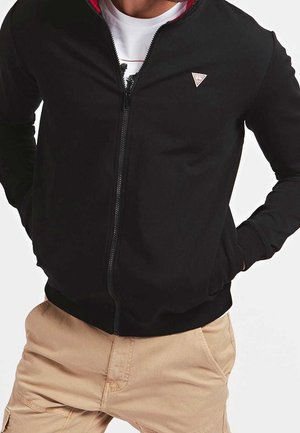 Mężczyzna noszący czarną kurtkę na zamek z małym trójkątnym logo, beżowe spodnie typu cargo oraz białą koszulkę z grafiką pod spodem.