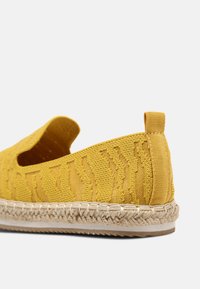 Geltonos espadrilės su tekstūruotu viršumi ir atvira šone. Yra džutiniu apklotu padu ir mažu užpakaliniu užtrauktuku, kad būtų patogu užsidėti.