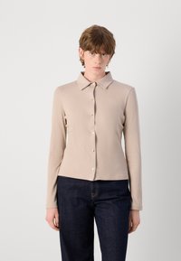 Beige Langarmhemd mit Button-Down-Kragen, gerippter Textur und goldfarbigen Knöpfen, kombiniert mit dunkelblauen Jeans.