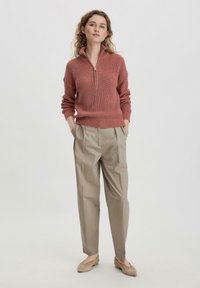Rostfarbener Strickpullover mit halbem Reißverschluss-Kragen, kombiniert mit hellbeigen, plissierten Hosen und taupefarbenen Slippern. Glatte Textur insgesamt.