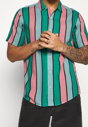 Mężczyzna w koszuli z krótkim rękawem zapinanej na guziki, z pionowymi paskami w kolorze zielonkawym, różowym i jasnoniebieskim, ze splecionymi rękami, pokazujący tatuaże na ramionach i palcach.