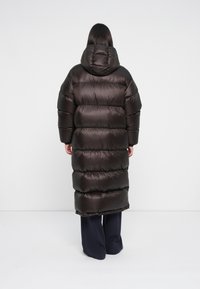 Persoon gekleed in een lange, zwarte, pufferjas met een capuchon, staand met de rug naar ons toe tegen een effen witte achtergrond.