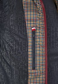 Windbreaker mit schwarzem Außenmaterial; das Innenfutter weist ein Karo-Muster in Braun, Blau und Beige auf; umfasst eine Reißverschlusstasche mit roter Akzentuierung.