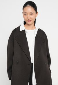 Abrigo de gran tamaño en marrón oscuro hecho de tela gruesa, con un cuello muesca, botones dobles y una textura suave, llevado sobre una camisa blanca.