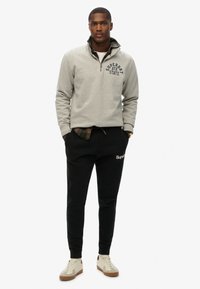 Lichtgrijze ziptrui met het "Superdry" logo, gedragen over een geblokt shirt. Zwarte joggers met "Superdry" tekst, gecombineerd met crèmekleurige sneakers.