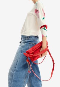 Kobieta w niebieskich dżinsach i białym swetrze w kwiaty trzyma czerwoną skórzaną torbę na ramię z dekoracyjnym plecionym paskiem i frędzlami.