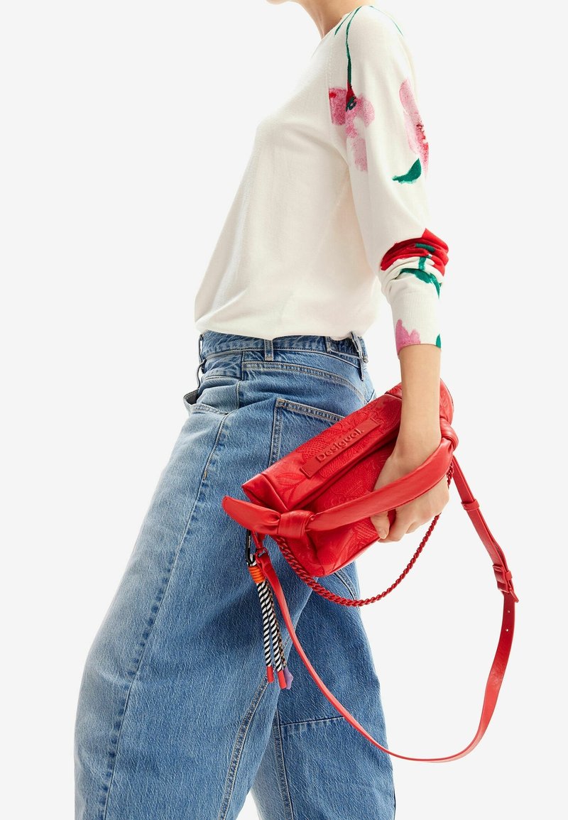 Kobieta w niebieskich dżinsach i białym swetrze w kwiaty trzyma czerwoną skórzaną torbę na ramię z dekoracyjnym plecionym paskiem i frędzlami.