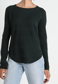 Mörkgrön stickad långärmad tröja med ett strukturerat randigt mönster, rund halsringning och rundad kant. Bärs med ljusblå jeans.